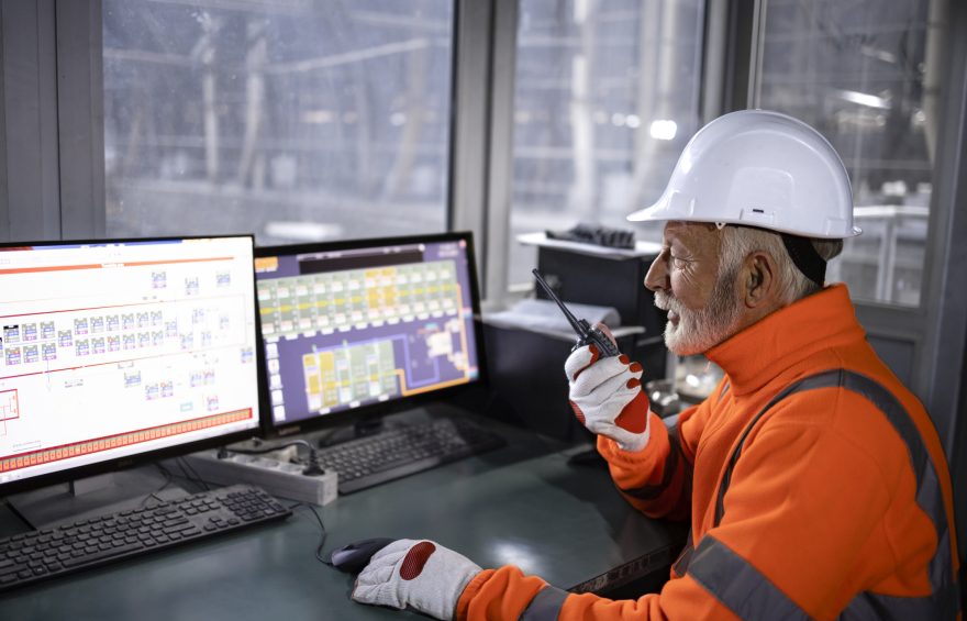 Industrial worker in safety equipment sitting in factory control room monitoring production process and talking on radio communication.